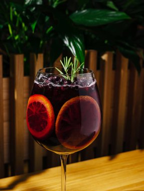Renkli bir içecek üzerinde kan portakalı dilimleri ve biberiye filizi bulunur. Arka planda yeşillik olan rahat bir ortamda ahşap bir masaya kurulur..