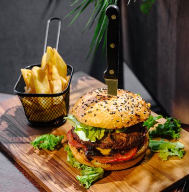 Susamlı çörek ile kalın bir hamburger marul, domates ve peynirle kaplıdır. Merkeze bir bıçak yerleştirilir. Küçük bir sepette kızarmış patates, ahşap bir yüzey üzerinde tabağa eşlik eder..