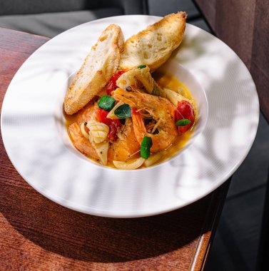 Karides, domates ve otlardan oluşan lezzetli bir deniz ürünleri tabağı, kızarmış ekmeğin yanında servis ediliyor. Yemek ahşap bir masaya konur ve yumuşak ışıklandırma sıcak bir ortam yaratır..