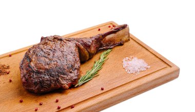 Büyük, sulu bir pirzola bifteği ahşap bir kesme tahtasının üzerinde duruyor. Bitkilerle çeşnilendirilir ve deniz tuzu eşlik eder. Biftek mükemmel kızartılmış, zengin rengini ve dokusunu gösteriyor..