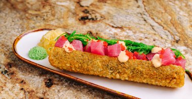 Dikkatlice hazırlanmış suşi rulosu taze ton balığı parçaları, deniz yosunu, wasabi ve turşulu zencefille süslenmiş. Tabak, kırsal bir zeminde dekoratif bir tabakta servis ediliyor..