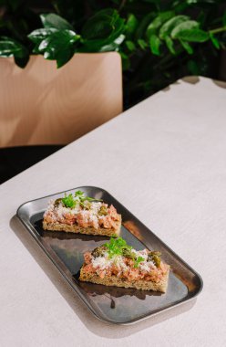 Açık yüzlü sandviçler lezzetli bir şekilde metalik tepside duruyor. Samimi atmosfer arka planda yeşil bitkiler barındırıyor ve sahneye tazelik katıyor..