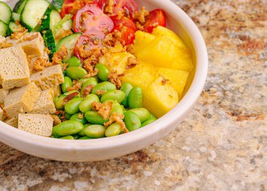 Salatalık, domates gibi taze sebzelerle dolu renkli bir kase ve ananas gibi meyvelerin yanı sıra tofu ve çıtır çıtır soslar. Sahne dokulu bir yüzey üzerinde canlı..