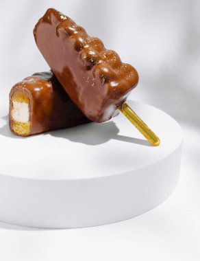 İki çikolatayla kaplı dondurma dairesel beyaz bir tabanda duruyor, yumuşak dokularını ve şekillerini yumuşak, bulanık bir zemine karşı sergiliyorlar. Odak, onların lezzetli görünümlerini vurguluyor..