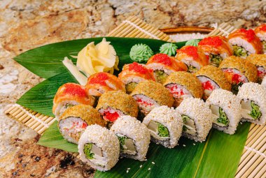 Renkli bir suşi tabağı ahşap bir tahtanın üzerinde çeşitli suşi ruloları sergiliyor. Garnitürler arasında zencefil ve kremalı yeşil soslar dilimleri ve taze dokunuşlar yer alıyor..