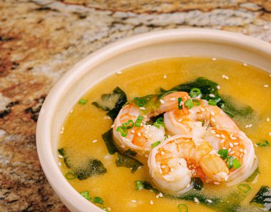 Baharatlı karides çorbası zengin bir çorbada taze karides ve taze yosun içerir. Yeşil soğan ve susamla süslenmiş granit bir yüzeye benziyor..