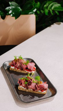 İki biftekli sandviç şık bir tepside otlarla süslenmiş olarak duruyor. Tablo minimalist bir tasarım sergiler ve yapraklı bir arkaplan ortama doğanın dokunuşunu ekler.