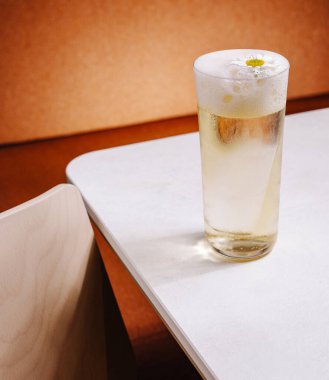 Hafif bir içecekle dolu berrak bir bardak pürüzsüz beyaz bir masada oturur. Narin bir çiçek en tepede süzülür, sıcak bir ortamda ferahlatıcı içeceğe biraz zarafet katar..