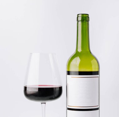 Bir bardak kırmızı şarap açılmamış bir şişenin yanında oturur. Şişenin basit bir etiketi var ve her ikisi de temiz, beyaz bir arka planda. Bu sahne bir rahatlama ya da kutlama anı öneriyor..