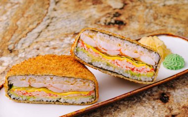 Bu yemekte ikiye bölünmüş çıtır bir suşi sandviçi, karides, avokado ve yumurta katmanları var. Renkli soslarla dokulu bir tabakta sunuluyor..