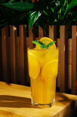 Canlı turuncu bir içecekle dolu uzun bir bardak ahşap bir masada oturur. Taze limon dilimleri ve nane yaprakları renk ve tazelik ekler, sıcak bir gün için canlandırıcı bir görüntü yaratır..