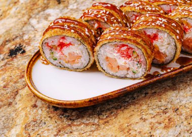 Renkli suşi ruloları dekoratif bir tabakta güzelce düzenlenir. Ekmeğe taze malzemeler ve üzerine susam tohumu serpiştirilmiş bir sos serpiştiriliyor..