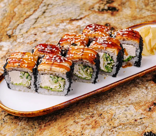 Taze suşi ruloları ahşap bir tabakta güzelce düzenlenir. Ekmekler kremalı ve parlak bir jöle ile kaplanmış, susam tohumları serpiştirilmiş ve limonla servis edilmiş..