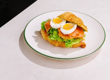 Füme somon, kıtır marul ve haşlanmış yumurta katmanlı taze kruvasan sandviçi. Parlak renkler ve dokular basit bir tabakta iştah açıcı bir sunum oluşturur.