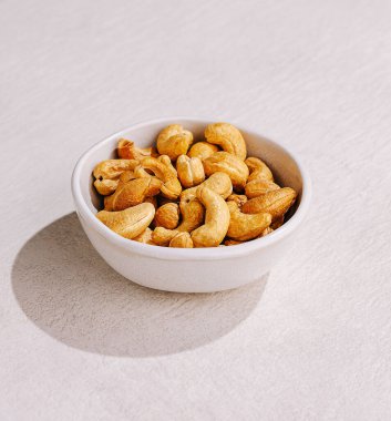 Kavrulmuş kaju fıstığıyla dolu küçük beyaz bir kase açık renkli bir yüzeye oturur. Cevizler altın sarısı ve hafif parlak. Lezzetli bir aperatif seçeneği ekliyor..