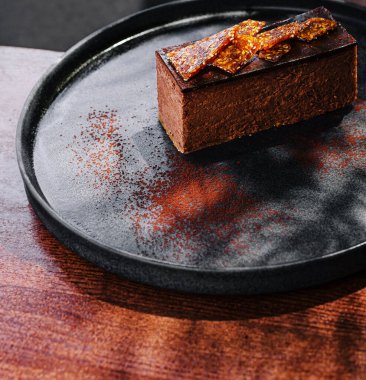 Zengin bir çikolatalı tatlı, üzerinde çıtır çıtır karamel parçaları olan siyah bir tabakta oturur. Dekorlar ahşap bir masa gösteriyor, tatlı bir ikram için davetkar bir atmosfer yaratıyor..