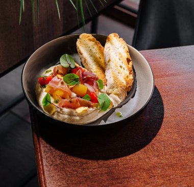 Özenle hazırlanmış bir tabakta, kremalı bir yatakta kesilmiş etler ve kesilmiş sebzeler, közlenmiş ekmekle sunuluyor, şık bir kafede ahşap bir masaya konuyor..