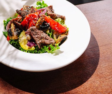 Taze yeşillikli canlı bir salata, ızgara biftek şeritleri, kiraz domatesleri, soğanlar ve mantarlar ahşap bir masada beyaz bir kasede zarif bir şekilde düzenlenir ve lezzetli bir yemek sergilenir..