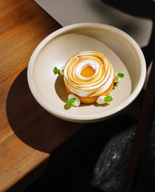 Zarif bir tatlı ahşap bir masanın üzerinde bej bir tabakta oturur. Tatlı, taze nane yapraklarıyla süslenmiş parlak sarısı ile sarmal kremalı bir üst içeriyor..