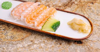 Güzel düzenlenmiş suşi tabağında salatalık dilimlerinin üzerinde taze karides bulunur. Wasabi ve turşulu zencefil tabağa eşlik ediyor. Rahatlatıcı bir yemek ya da özel bir durum için mükemmel..