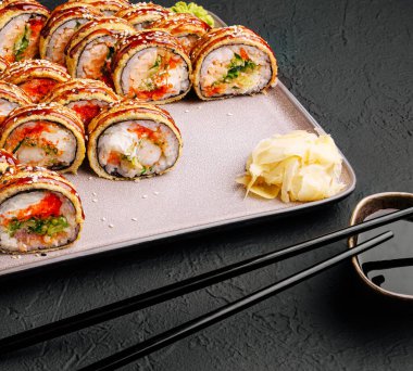 Taze suşi ruloları, zencefil turşusuyla süslenmiş ve küçük bir tabak soya sosuyla servis edilmiş bir tabakta güzelce düzenlenir. Zarif yemek çubukları sahneyi tamamlıyor..