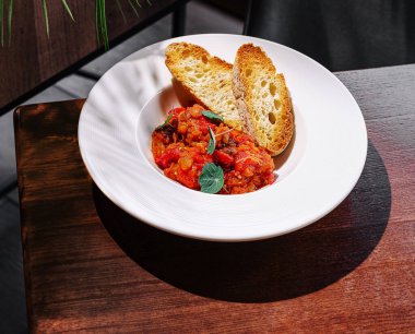 Bitkilerle bezenmiş domates soslu canlı bir yemek beyaz bir kasede oturur. Artistik olarak yanına iki parça kızarmış ekmek konur. Ortam sıcak ve davetkar..