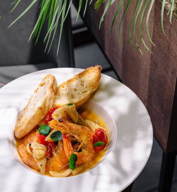 Canlı bir deniz ürünleri tabağında karides ve sebze, yanında da kızarmış ekmek bulunur. Güneşli bir masada gölgeli bitki yapraklarıyla güzelce sunuluyor..