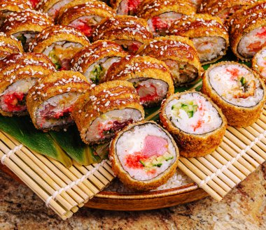 Çeşitli suşi dürümleri ahşap bir tabağa yerleştirilmiş ve altında bambu hasırı var. Renkli rulo farklı malzemeler ve dokular içeriyor, cazip bir ekran oluşturuyor..