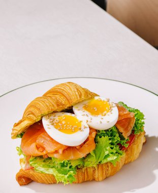 Kruvasan sandviçi füme somon, taze marul, dilimlenmiş yumurta ve susam tohumuyla doludur. Bu yemek, beyaz tabakta servis edilen kahvaltı ya da brunch için mükemmel..