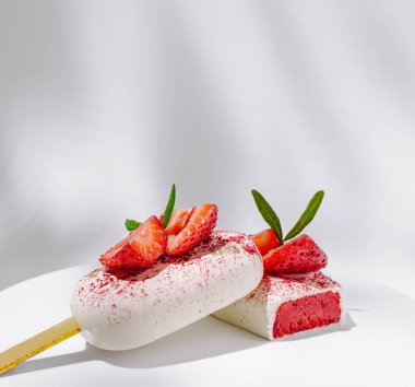 İki cazip çilekli tatlı beyaz bir tabakta oturur. Biri dilimlenmiş çileklerle kaplı kremalı dondurma, diğeri ise kare şeklinde, meyveli lezzet ve dekorasyonla dolu..