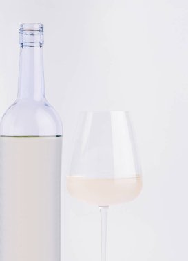 Beyaz şarapla dolu berrak bir bardak şeffaf bir şişenin yanında durur. Yumuşak ışıklandırma şarabın rengini vurgular, zarif ve basit bir görüntü yaratır..