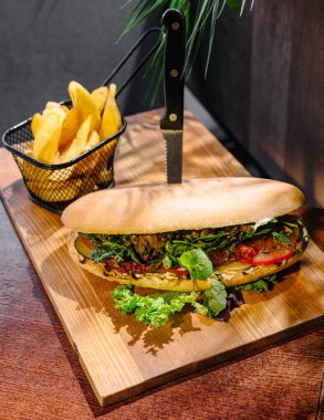 Sebze ve otlarla dolu taze yapılmış sandviç ahşap bir tahtanın üzerinde, kızarmış patates sepetinin yanında, sıcak bir ortamda canlı bir yemek yakalıyor..