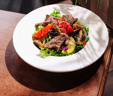 Izgara biftekli canlı bir salata, renkli biberler ve çeşitli sebzeler beyaz bir tabakta oturur. Tabak, rahat bir restoranda, ahşap bir masaya konur..