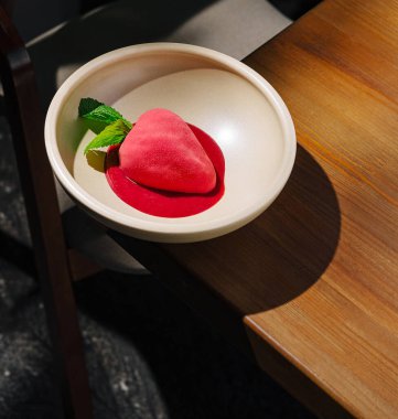 Güzel bir tatlı, zengin bir kırmızı sosun üzerinde duran çilek şeklinde pembe bir şerbet içerir. Taze nane yaprakları renk katar. Bu sanatsal yemek, rahat bir kafedeki ahşap bir masaya kurulmuş..