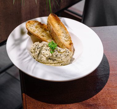 Lezzetli kremalı bir sos, yanında iki dilim kızarmış ekmek ile birlikte beyaz bir kasede servis edilir. Ayar, sıcak ışıklandırma ve ahşap dokular içeriyor ve hoş bir atmosfer yaratıyor..