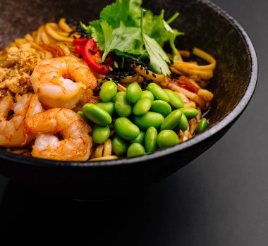 Karides, edamame ve taze otlarla dolu canlı bir kase siyah bir yüzeye oturur. Bu yemek renkli şehriyeleri sergiliyor. Lezzetli ve dengeli bir yemek sunuyor. Öğle veya akşam yemeği için..