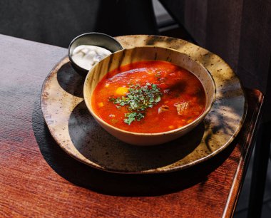 Sıcak bir kase doyurucu çorba kırsal bir ahşap masada oturur. Canlı kırmızı çorba taze otlarla kaplıdır ve yanında küçük bir kremalı ekşi krema ile rahatlatıcı bir yemek sunulur..