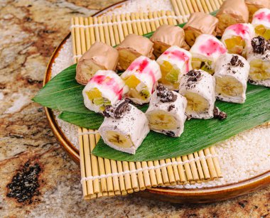 Çeşitli tatlı suşi dürümleri ahşap bir tabakta düzgünce düzenlenir. Ekmekler renkli malzemeler içeriyor ve pilavın üzerine bambu bir hasırın üzerine konuyor. Ayar, sıradan bir yemek atmosferini çağrıştırıyor..