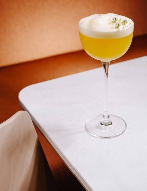 Serinletici sarı bir kokteyl modern bir masadaki saplı bir bardakta zarif bir şekilde duruyor. Köpüklü ve süslü içecekler sunar. Keyifli bir ortam sunar..