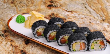 Siyah suşi ruloları beyaz bir tabakta düzgünce düzenlenir. Yeşil wasabi ve turşulu zencefil eşliğinde, bu aşçılık yaratımı canlı ve davetkar..