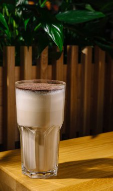 Uzun bir bardak buzlu kahve, üzerinde çikolata tozu olan ahşap bir masanın üzerinde oturur. Bereketli yeşil bitkiler canlı bir zemin oluşturarak canlandırıcı bir atmosfer yaratırlar..