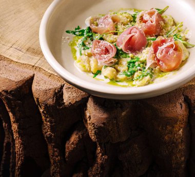 Kırsal ahşap bir masada servis edilen taze yeşillik ve prosciutto ile güzel düzenlenmiş bir yemek. Hassas sunum yemeğin canlı renklerini ve dokularını vurguluyor..