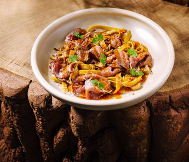 Çarpıcı bir makarna tabağında yumuşak et şeritleri, zengin soslar ve taze otlar bulunur. Tabak kaba ahşap bir yüzeye yerleştiriliyor ve sunuma sıcaklık ve doku katıyor..