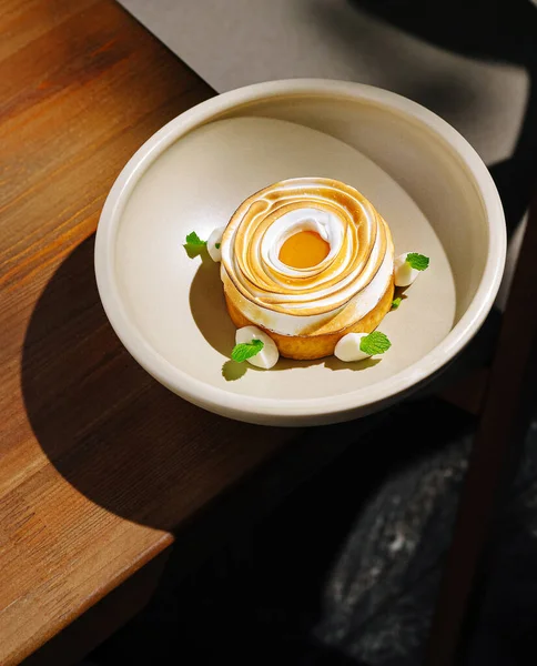 Zarif bir tatlı ahşap bir masanın üzerinde bej bir tabakta oturur. Tatlı, taze nane yapraklarıyla süslenmiş parlak sarısı ile sarmal kremalı bir üst içeriyor..