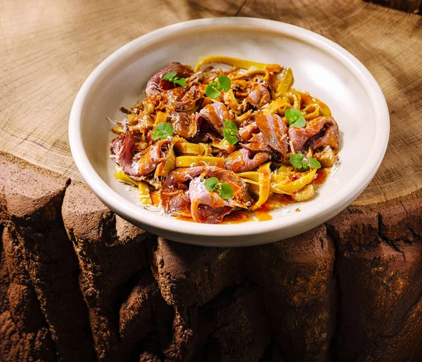 Çarpıcı bir makarna tabağında yumuşak et şeritleri, zengin soslar ve taze otlar bulunur. Tabak kaba ahşap bir yüzeye yerleştiriliyor ve sunuma sıcaklık ve doku katıyor..