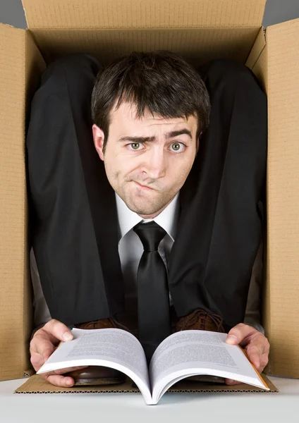 Homem de negócios flexível com rosto duvidoso em uma caixa de papelão lendo um livro — Fotografia de Stock