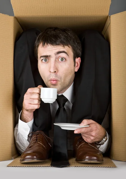 Flexible businessman with doubtful face in a cardboard box drinking coffee — Stock Photo, Image