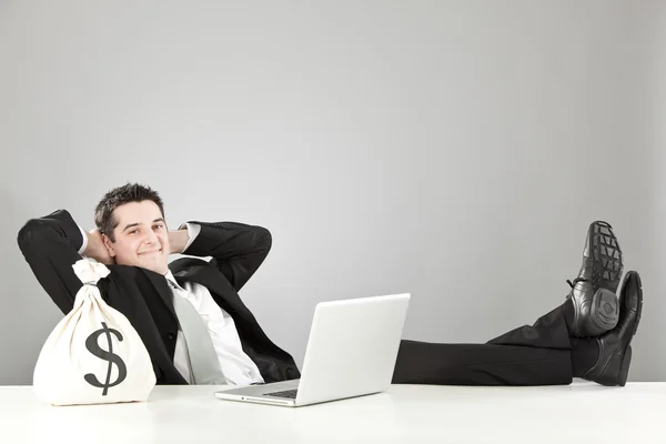 Homem de negócios feliz relaxado com saco de dinheiro e laptop isolado em cinza — Fotografia de Stock