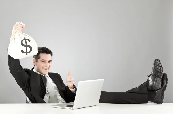 Homem de negócios feliz relaxado com saco de dinheiro e laptop isolado em cinza — Fotografia de Stock