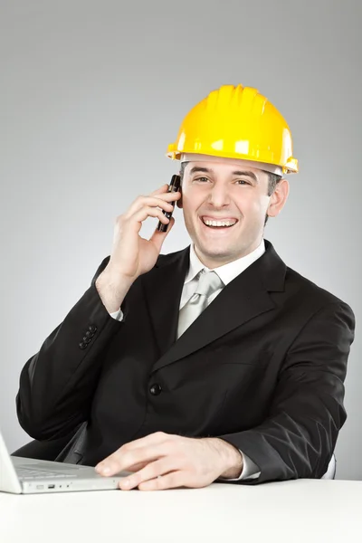 Schön lächelnder Ingenieur mit gelbem Helm und Laptop, der isoliert auf grau telefoniert — Stockfoto